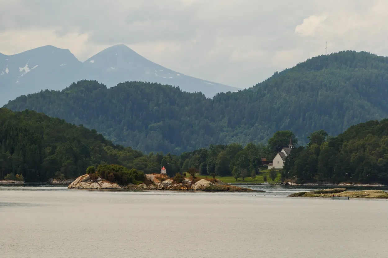 Small Norwegian church among trees beside a calm fjord with distant mountains