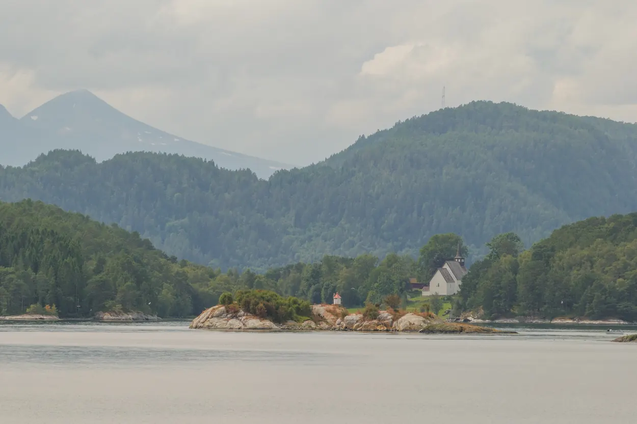 Distant church and rocky island framed by forested mountains across a wide fjord