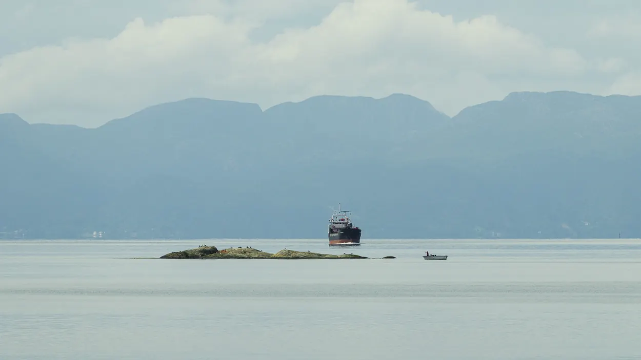 Cargo ship and a small boat near a rocky islet on a misty blue fjord